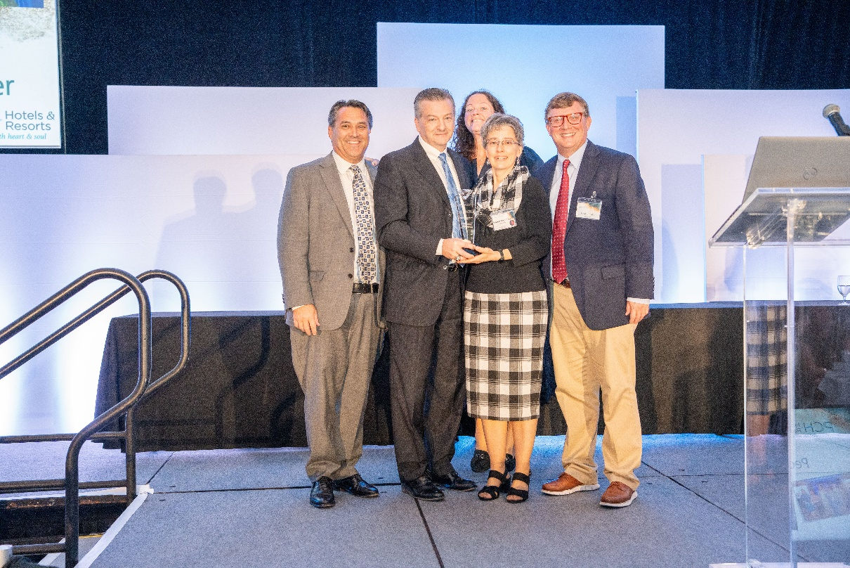 Karen Moulton receives the 2025 Bucky Miller Award from PCH Hotels & Resorts President, Tony Davis, joined by leaders from the Renaissance Riverview Plaza Hotel.